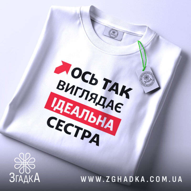 Футболка ось так виглядає ідеальна сестра. Біла футболка, складена на столі, з видимим принтом. На фоні світлий однотонний колір. Принт містить червоний текст на чорному фоні, що говорить: 'Ось так виглядає ідеальна сестра', а також зелена етикетка з логотипом.