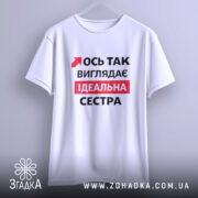 Футболка ось так виглядає ідеальна сестра. Біла футболка, підвішена на вішалці, з коротким рукавом. На фоні м'який сірий колір. Принт на футболці містить червоний текст з чорними елементами, що говорить: 'Ось так виглядає ідеальна сестра'.