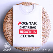 Футболка ось так виглядає ідеальна сестра. Біла футболка, складена на плетеній кошику, з видимим принтом. На фоні світлий однотонний колір. Принт містить червоний текст на чорному фоні, що говорить: 'Ось так виглядає ідеальна сестра'.