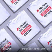 Футболка ось так виглядає ідеальна сестра. Біла футболка з коротким рукавом, зображена на моделі, яка позує з легким усмішкою. На фоні м'який пастельний колір. Принт на футболці містить червоний текст з чорними елементами, що говорить: 'Ось так виглядає ідеальна сестра'.