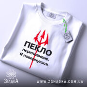 Футболка пекло переповнене, біла футболка складена на поверхні, зображена зверху, фон світло-сірий, принт червоний тризуб та чорний текст, з етикеткою зеленого кольору.