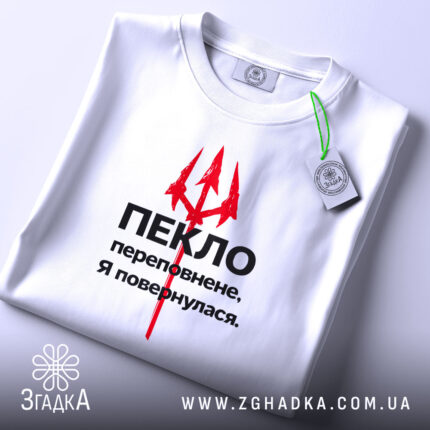 Футболка пекло переповнене, біла футболка складена на поверхні, зображена зверху, фон світло-сірий, принт червоний тризуб та чорний текст, з етикеткою зеленого кольору.