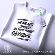 Футболка підхід до мене. Складена біла футболка з чорним принтом, з етикеткою зеленого кольору, на фоні світло-сірого кольору.