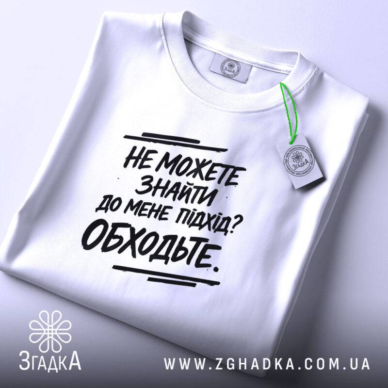 Футболка підхід до мене. Складена біла футболка з чорним принтом, з етикеткою зеленого кольору, на фоні світло-сірого кольору.