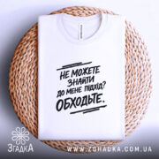 Футболка підхід до мене. Складена біла футболка з чорним принтом, розташована на плетеній кошику, на фоні світло-сірого кольору.