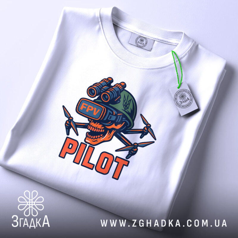 Футболка Пілот FPV білого кольору, складена на поверхні, з видимим принтом черепа з окулярами віртуальної реальності та дронами. Принт яскравий, з червоними, зеленими та чорними елементами. На футболці також видно етикетку з логотипом бренду, що підкреслює якість матеріалу.