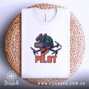 Футболка Пілот FPV білого кольору, складена на плетеній кошику, з яскравим принтом черепа в окулярах віртуальної реальності та дронами. Принт виконаний в червоних, зелених та чорних кольорах, а фон м'який та нейтральний, що підкреслює текстуру тканини.