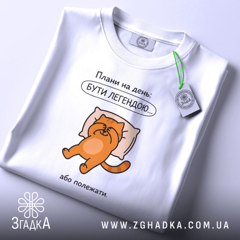 Футболка плани на день: біла футболка, зображення оранжевого кота на подушці, текст "Плани на день: БУТИ ЛЕГЕНДОЮ...". Футболка лежить на світлому фоні, видно етикетку з зеленим шнурком.