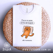 Футболка плани на день: біла футболка, зображення оранжевого кота на подушці, текст "Плани на день: БУТИ ЛЕГЕНДОЮ...". Футболка лежить на плетеній основі, видно етикетку.