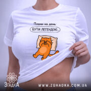 Футболка плани на день: біла футболка, зображення оранжевого кота на подушці, текст "Плани на день: БУТИ ЛЕГЕНДОЮ...". Футболка лежить на світлому фоні, видно етикетку з зеленим шнурком.