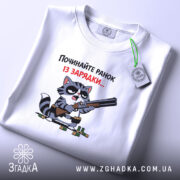 Футболка починайте ранок із зарядки. Біла футболка, складена на столі, з принтом кота з рушницею. Кот має чорні окуляри, фон світло-сірий. Текст на футболці червоний і чорний, етикетка з зеленим шнурком.