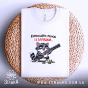 Футболка починайте ранок із зарядки. Біла футболка, складена на плетеному кошику, з принтом кота з рушницею. Кот має чорні окуляри, фон білий. Текст на футболці червоний і чорний.