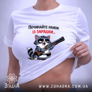 Футболка починайте ранок із зарядки. Біла футболка, складена на столі, з принтом кота з рушницею. Кот має чорні окуляри, фон світло-сірий. Текст на футболці червоний і чорний, етикетка з зеленим шнурком.