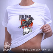 Футболка погляд вічності. Біла футболка, складена на столі, з принтом чорного ворона на черепі, оточеного червоними графічними елементами. Вид зверху, з етикеткою, що звисає з коміра.