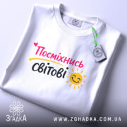 Футболка посміхнись світові, біла, складена на поверхні, фон світлий, принт з написом 'Посміхнісь світові' у яскравих кольорах, з етикеткою на комірці.