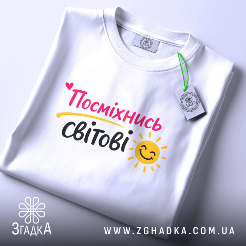 Футболка посміхнись світові, біла, складена на поверхні, фон світлий, принт з написом 'Посміхнісь світові' у яскравих кольорах, з етикеткою на комірці.