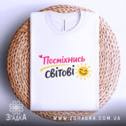 Футболка посміхнись світові, біла, складена на плетеній основі, фон світлий, принт з написом 'Посміхнісь світові' у яскравих кольорах, з усмішкою та сонечком.