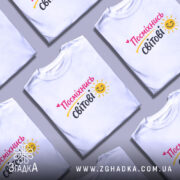 Футболка посміхнись світові, біла, зображена на моделі з легким ракурсом, фон пастельного кольору, принт з написом 'Посміхнісь світові' у яскравих кольорах з усмішкою та сонечком.