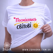 Футболка посміхнись світові, біла, складена на поверхні, фон світлий, принт з написом 'Посміхнісь світові' у яскравих кольорах, з етикеткою на комірці.