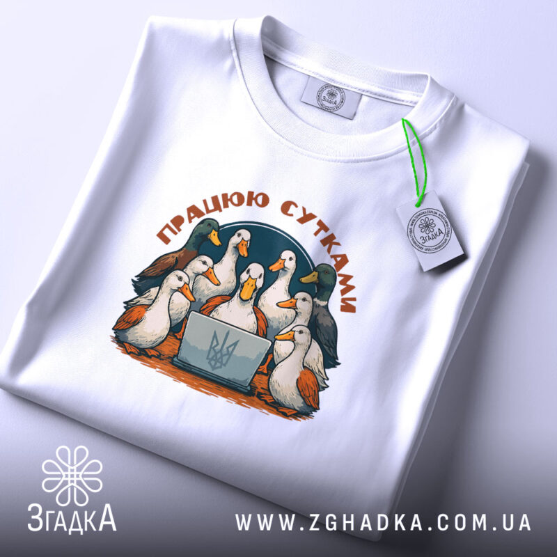 Футболка працюю сутками, біла, зображена на столі, ракурс зверху, фон світло-фіолетовий, принт з птахами навколо ноутбука, з написом 'ПРАЦЮЮ СУТКАМИ', на етикетці зелена стрічка.