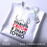 Футболка правлять світом, біла, зображена на столі, ракурс зверху, фон світло-сірий, принт з написом 'Правлять світом я знаю точно' у червоному та чорному кольорах, з малюнком грудей, з етикеткою на шнурку.
