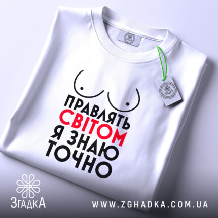 Футболка правлять світом, біла, зображена на столі, ракурс зверху, фон світло-сірий, принт з написом 'Правлять світом я знаю точно' у червоному та чорному кольорах, з малюнком грудей, з етикеткою на шнурку.