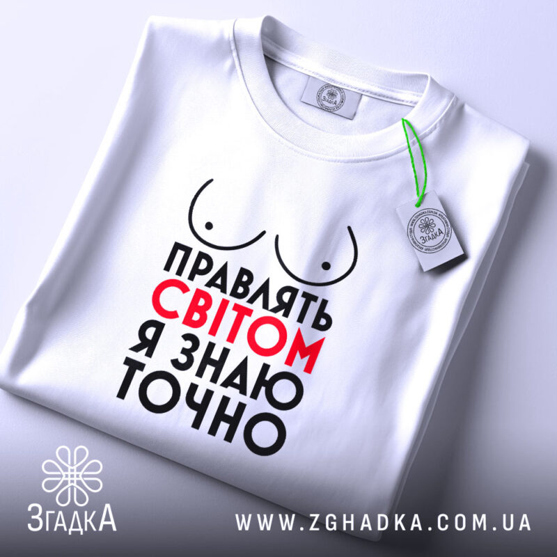 Футболка правлять світом, біла, зображена на столі, ракурс зверху, фон світло-сірий, принт з написом 'Правлять світом я знаю точно' у червоному та чорному кольорах, з малюнком грудей, з етикеткою на шнурку.