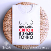 Футболка правлять світом, біла, зображена на плоскій поверхні, фон світло-сірий, принт з написом 'Правлять світом я знаю точно' у червоному та чорному кольорах, з малюнком грудей, розташована на плетеному кошику.