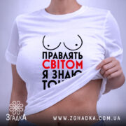 Футболка правлять світом, біла, зображена на столі, ракурс зверху, фон світло-сірий, принт з написом 'Правлять світом я знаю точно' у червоному та чорному кольорах, з малюнком грудей, з етикеткою на шнурку.