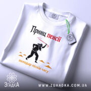 Футболка принц пенсії, біла, лежить на світлому фоні, принт з чорним силуетом персонажа з червоною палицею, текст 'Принц пенсії' у верхній частині, видна етикетка з зеленим шнурком.