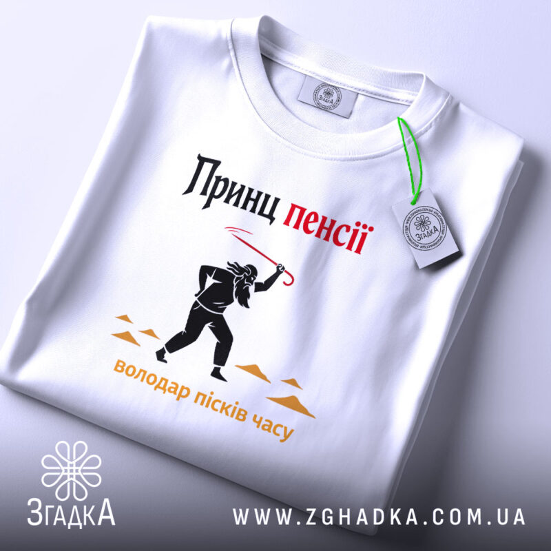 Футболка принц пенсії, біла, лежить на світлому фоні, принт з чорним силуетом персонажа з червоною палицею, текст 'Принц пенсії' у верхній частині, видна етикетка з зеленим шнурком.