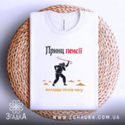 Футболка принц пенсії, біла, лежить на плетеній основі, принт з чорним силуетом персонажа з червоною палицею, текст 'Принц пенсії' у верхній частині, видно етикетку.