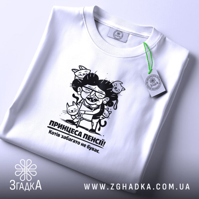 Футболка принцеса пенсії, біла, на столі, з верхнього ракурсу, фон світло-сірий, принт з карикатурою жінки з котами, текст під принтом: 'Котів забагато не буває.'