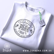 Футболка ручна робота з любов’ю, біла, складена на поверхні, фон світло-сірий, принт чорного кольору з текстом та квітковими елементами, на комірці видна етикетка.