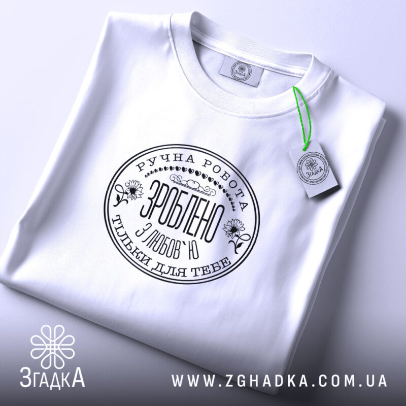 Футболка ручна робота з любов’ю, біла, складена на поверхні, фон світло-сірий, принт чорного кольору з текстом та квітковими елементами, на комірці видна етикетка.