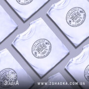 Футболка ручна робота з любов’ю, біла, на моделі з легким усмішкою, фон світло-фіолетовий, принт чорного кольору з текстом та квітковими елементами.