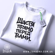 Футболка щастя прямо перед вами, біла, складена на поверхні, фон світло-фіолетовий, принт чорний з написом 'Щастя прямо перед вами', з етикеткою зеленого кольору.