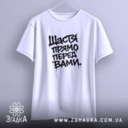 Футболка щастя прямо перед вами, біла, підвішена на вішалці, фон світло-сірий, принт чорний з написом 'Щастя прямо перед вами', з широкими рукавами.