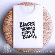 Футболка щастя прямо перед вами, біла, складена на плетеній основі, фон світло-фіолетовий, принт чорний з написом 'Щастя прямо перед вами'.