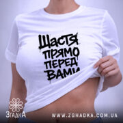 Футболка щастя прямо перед вами, біла, складена на поверхні, фон світло-фіолетовий, принт чорний з написом 'Щастя прямо перед вами', з етикеткою зеленого кольору.