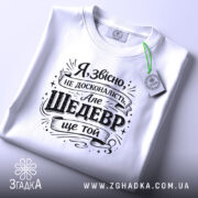 Футболка шедевр ще той. Біла футболка, складена на поверхні, з чорним принтом, фон світло-сірий, на футболці видно етикетку з зеленим шнурком.