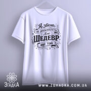 Футболка шедевр ще той. Біла футболка, підвішена на вішалці, фон світло-сірий, принт чорного кольору з текстом, що містить жартівливе висловлювання.