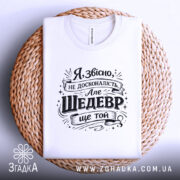 Футболка шедевр ще той. Біла футболка, складена на плетеній основі, фон білий, принт чорного кольору з текстом, що виглядає стильно і сучасно.