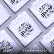 Футболка шедевр ще той. Біла футболка з чорним принтом, зображена в ракурсі з близької відстані, фон ніжно-фіолетовий, принт містить текст з кумедним висловом.