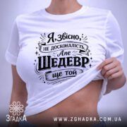 Футболка шедевр ще той. Біла футболка, складена на поверхні, з чорним принтом, фон світло-сірий, на футболці видно етикетку з зеленим шнурком.