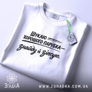 Футболка шукаю парубка, біла, знята зверху на плоскій поверхні, принт чорного кольору з текстом, що говорить про пошук хорошого парубка, з зеленим ярликом, що підкреслює стиль.