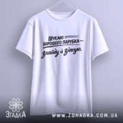 Футболка шукаю парубка, біла, підвішена на вішалці, з нейтральним фоном, принт чорного кольору з текстом, що говорить про пошук хорошого парубка, з широкими рукавами.