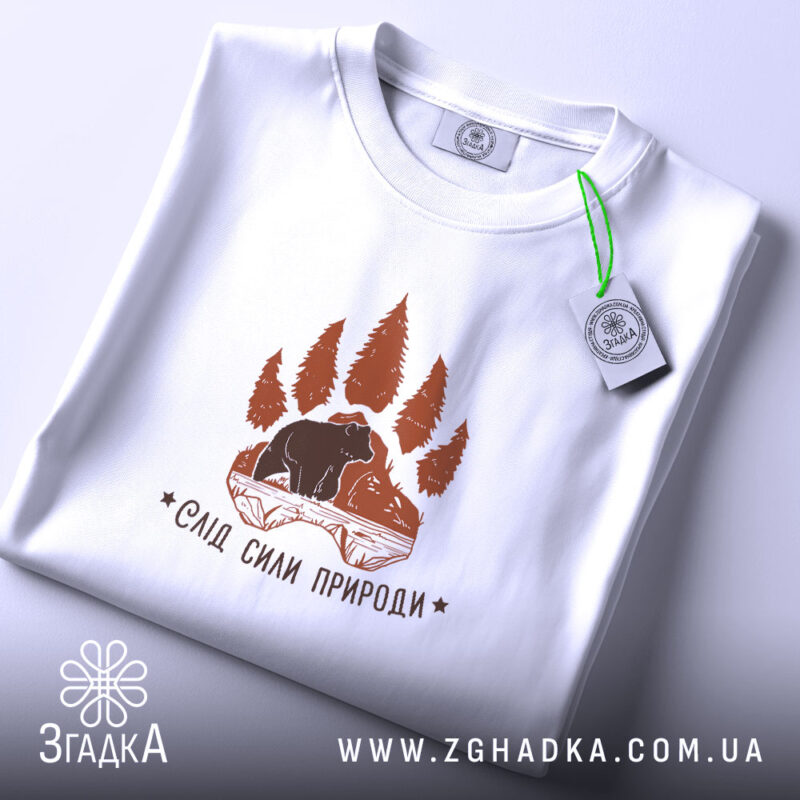 Футболка слід сили природи, біла, на плоскій поверхні, з етикеткою, фон світлий, принт коричневого кольору з ведмедем та деревами, вид зверху.