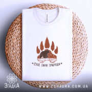 Футболка слід сили природи, біла, на плоскій поверхні, розташована на плетеній основі, фон світлий, принт коричневого кольору з ведмедем та деревами.