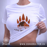 Футболка слід сили природи, біла, на плоскій поверхні, з етикеткою, фон світлий, принт коричневого кольору з ведмедем та деревами, вид зверху.